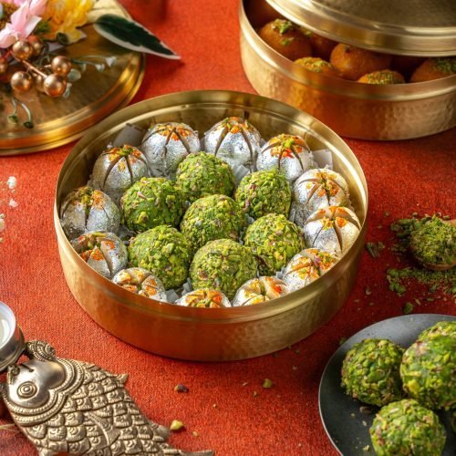 Brass round box filled with pistachio and silver-coated Indian laddus, placed on a festive red cloth with traditional decor.