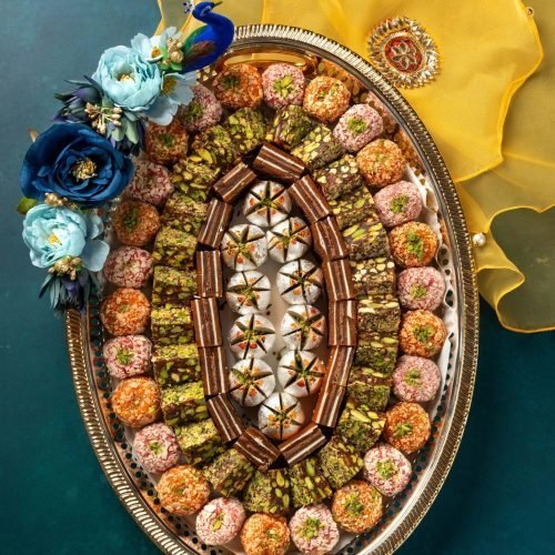 Decorative silver tray with an assortment of colorful Indian sweets, adorned with flowers and festive yellow cloth on the side
